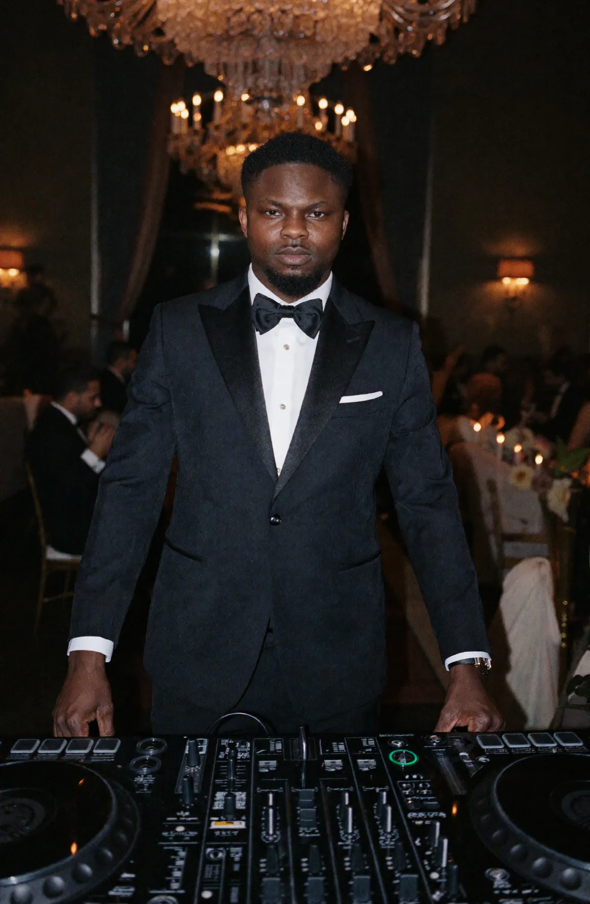 Ade Ajao, DJ Wrexx, in a black tuxedo standing behind DJ decks at an event